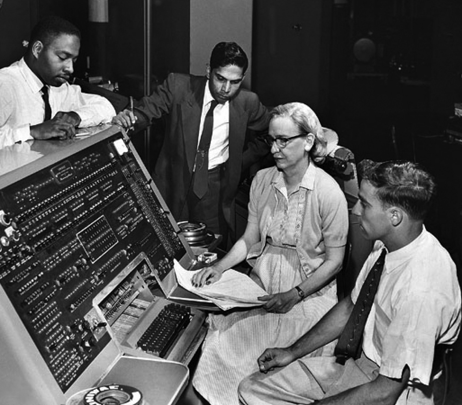 Grace Hopper, en uniforme de la marine américaine, devant les armoires de l'ordinateur UNIVAC, manipulant un panneau de commandes.