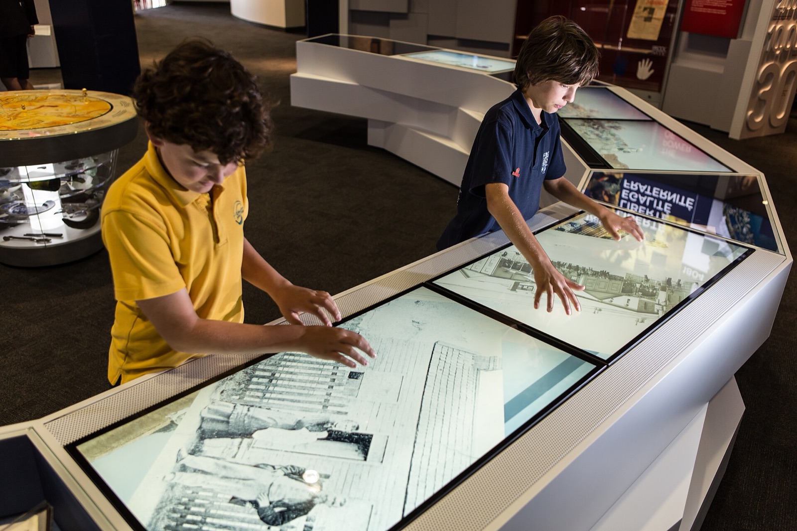 Deux enfants posent les mains sur les écrans tactiles inclinés d'un meuble de médiation dans une salle d'exposition ; les dalles affichent des photographies anciennes, et d'autres vitrines éclairées occupent l'arrière-plan.