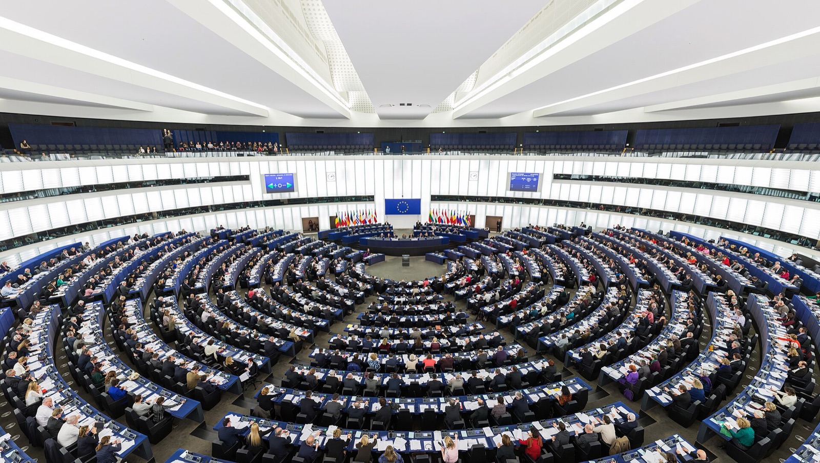 Vue plongeante de l'hémicycle du Parlement européen à Strasbourg en séance plénière : les eurodéputés sont assis aux pupitres bleus disposés en demi-cercles concentriques, dossiers ouverts devant eux, face à la tribune surmontée du drapeau européen et encadrée par les drapeaux des États membres, deux écrans affichant le décompte d'un vote.