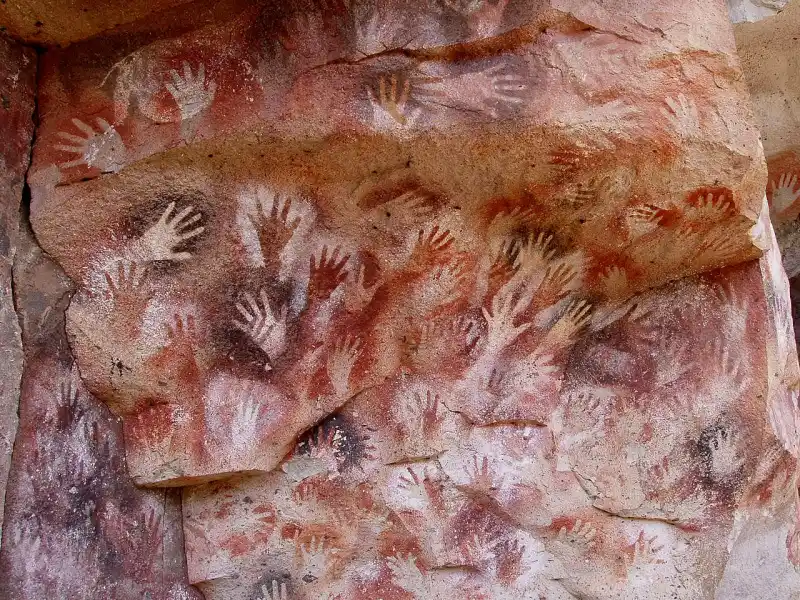 Paroi rocheuse ocre couverte de dizaines de mains en négatif, obtenues en projetant du pigment autour d&rsquo;une main appliquée contre la roche. Les empreintes se superposent par endroits et déclinent plusieurs teintes, du blanc au rouge sombre en passant par l&rsquo;ocre.