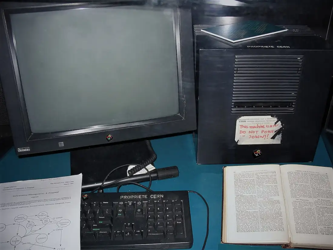Poste de travail NeXT exposé en vitrine : un écran cathodique noir, une unité centrale cubique portant une étiquette manuscrite « This machine is a server, DO NOT POWER IT DOWN!! », un clavier gravé « Propriété CERN », et posé devant, un tirage papier de la proposition d&rsquo;origine ouvert sur son schéma.