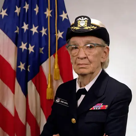 Portrait officiel de Grace Hopper en uniforme de la marine américaine, devant un drapeau des États-Unis
